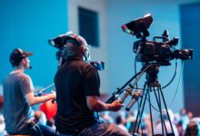 Two cameramen operate professional video cameras at an indoor event with a lively audience.