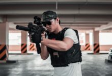 A man dressed in tactical gear aims a rifle in a shaded parking area, suggesting a training scenario.