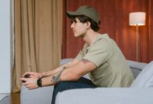 Tattooed man wearing cap playing video games on a sofa indoors.