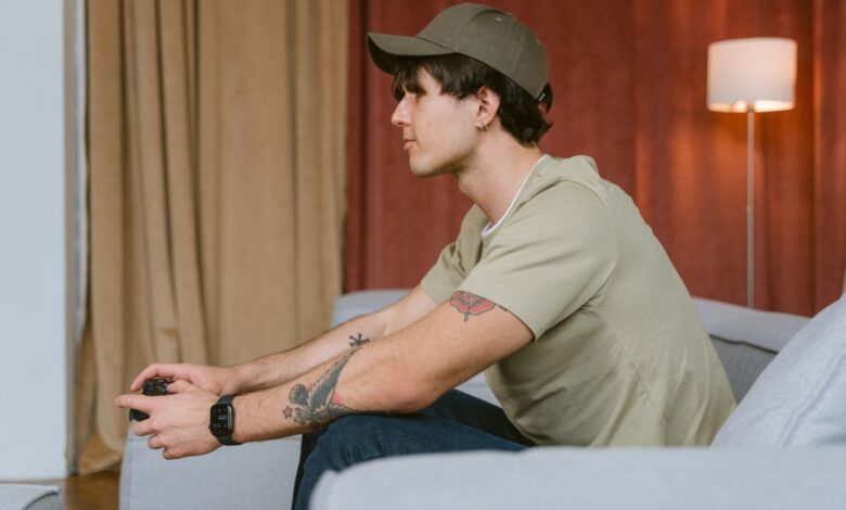 Tattooed man wearing cap playing video games on a sofa indoors.