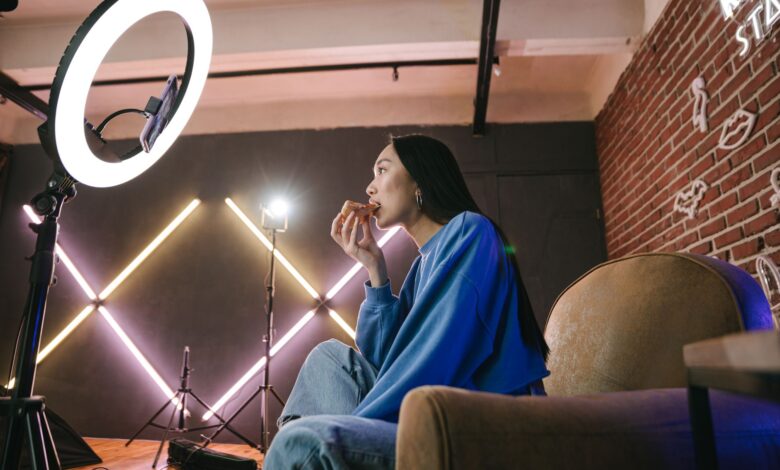 Asian woman content creator eating pizza while recording a video indoors with ring light.