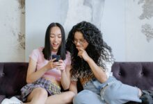 Two teenage girls happily sharing a moment on a sofa while looking at a smartphone indoors.