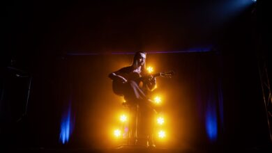 A musician playing an acoustic guitar on stage, illuminated by dramatic yellow and blue lights.