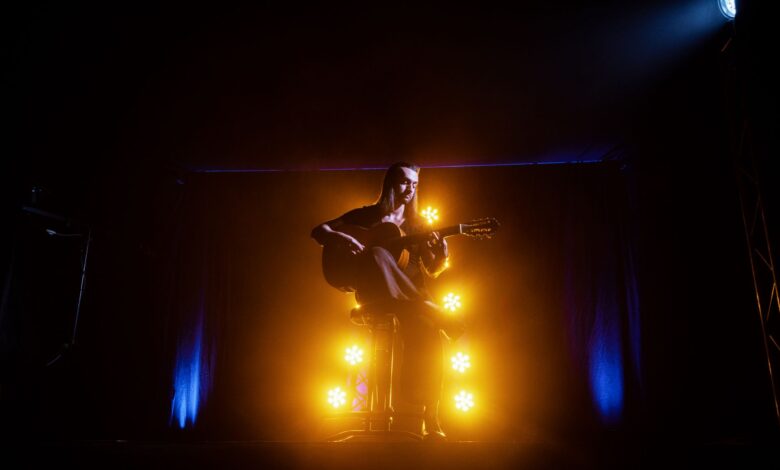 A musician playing an acoustic guitar on stage, illuminated by dramatic yellow and blue lights.
