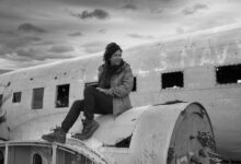 Black and white photo of a woman sitting on an abandoned plane in Iceland under a cloudy sky.
