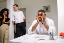 A stressed employee sits at a desk with colleagues in discussion nearby, highlighting workplace tension.