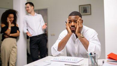 A stressed employee sits at a desk with colleagues in discussion nearby, highlighting workplace tension.