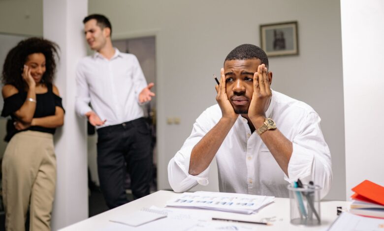 A stressed employee sits at a desk with colleagues in discussion nearby, highlighting workplace tension.