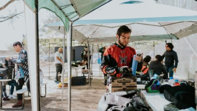 Group of adult men preparing gear for paintball under outdoor tents in daytime.