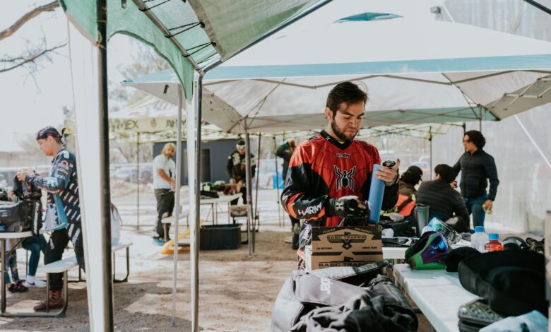 Group of adult men preparing gear for paintball under outdoor tents in daytime.