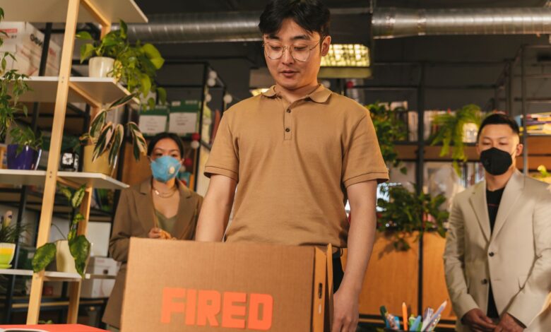 Man standing by cardboard box labeled 'fired', in an office surrounded by colleagues.