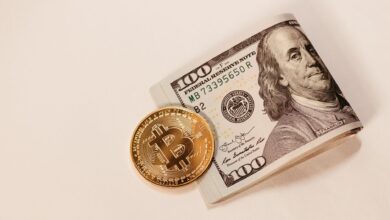 A Bitcoin coin placed next to a rolled US hundred dollar bill, symbolizing cryptocurrency and traditional currency.