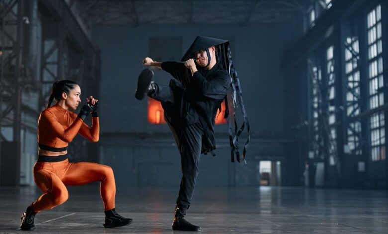 Two cosplayers performing martial arts poses in an industrial space, showcasing vibrant costumes and action.