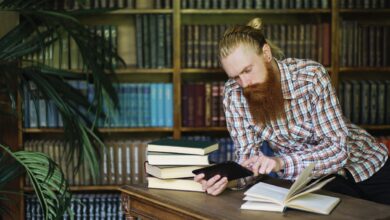 Bearded man reading a book and using a tablet in a classic library setting.