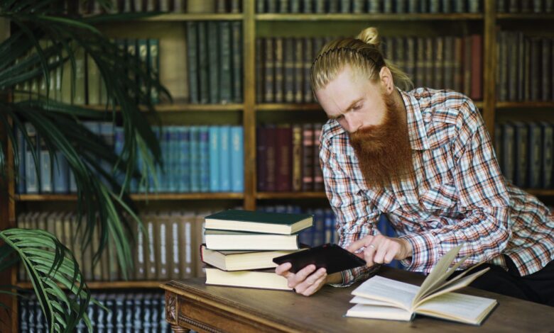 Bearded man reading a book and using a tablet in a classic library setting.