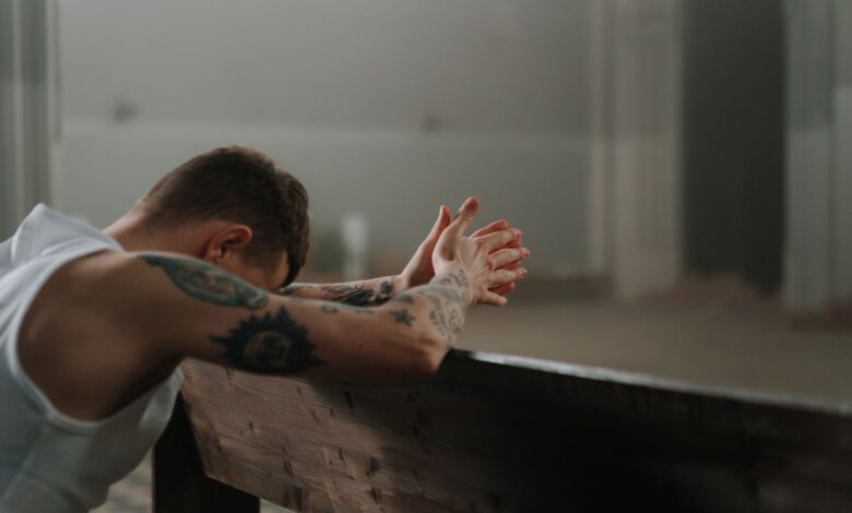 A tattooed man is solemnly praying indoors, reflecting spirituality and repentance.