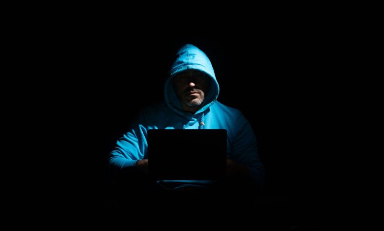 A person in a hoodie using a laptop in a dark setting, conveying mystery and technology themes.