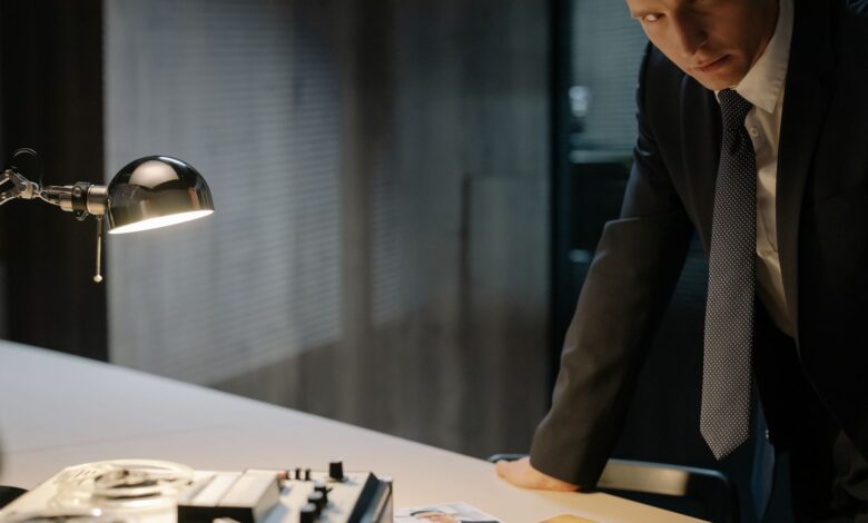 A detective in a suit examines evidence on a table under a lamp, focused on solving a case.