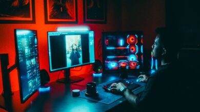 A young man working on a multi-screen computer setup in a dark, neon-lit room.