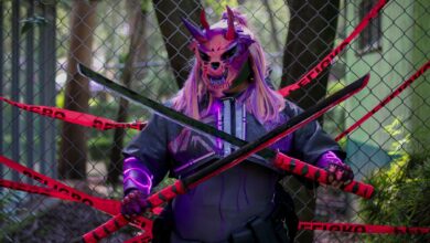 A person in a demon mask holds swords in front of a fence marked with warning tape.