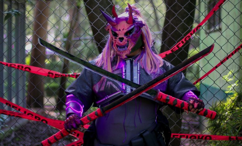 A person in a demon mask holds swords in front of a fence marked with warning tape.