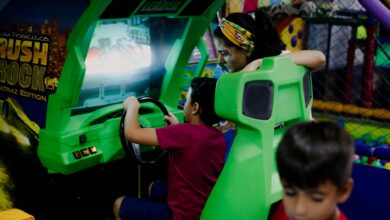 Unrecognizable child having fun with bright car racing simulator while turning steering wheel near serious ethnic woman and funny brother in shopping center near playground