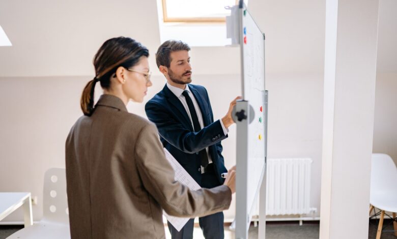 Two colleagues in formal attire discussing strategies on a whiteboard in a modern office space.
