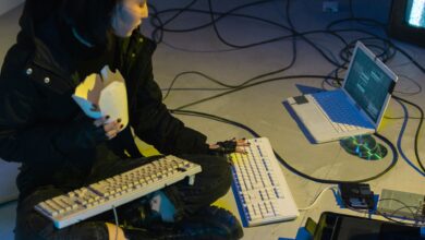 A young woman in a dark room surrounded by computers and cables, eating and typing on keyboards.