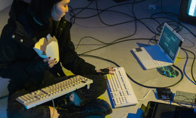 A young woman in a dark room surrounded by computers and cables, eating and typing on keyboards.