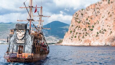Wooden pirate boat with sailing mast and painted skull floating on rippling sea near rocky mountain against grassy hilly area