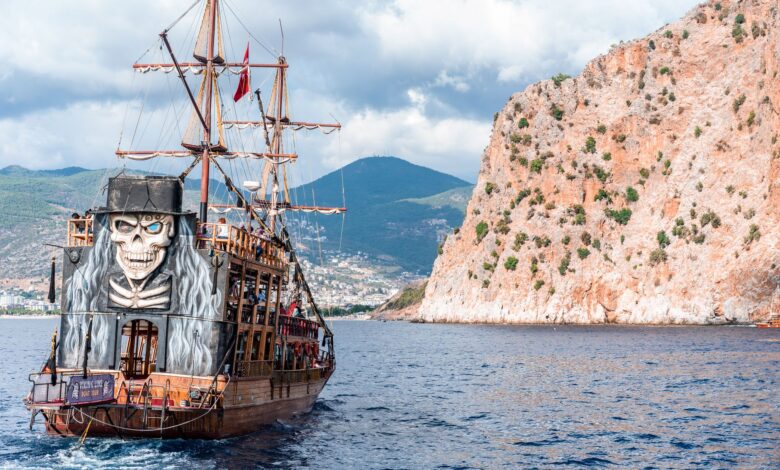 Wooden pirate boat with sailing mast and painted skull floating on rippling sea near rocky mountain against grassy hilly area