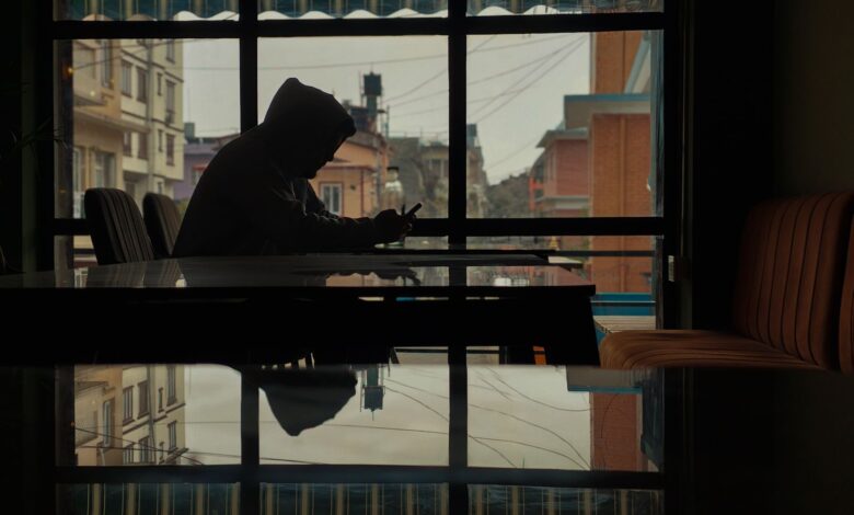 Silhouette of a person in a hoodie using a smartphone inside a café with street view.