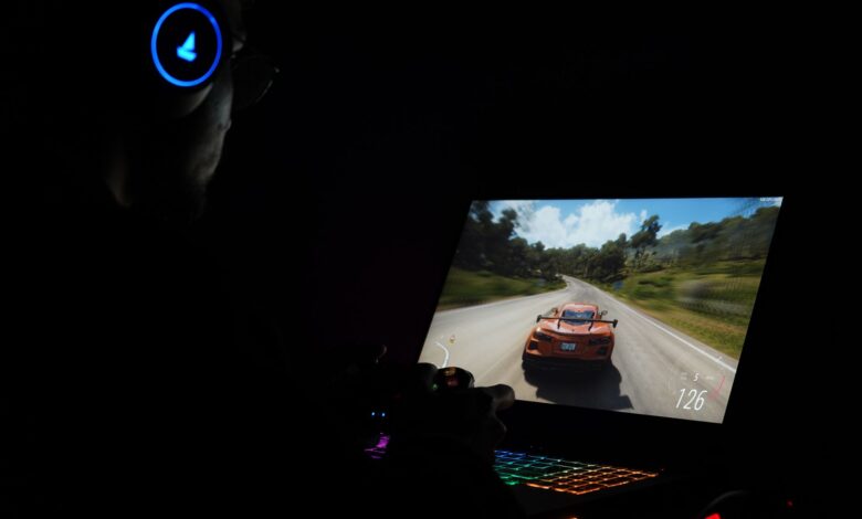 A gamer using a laptop and headphones to play a car racing game in a dark setting.