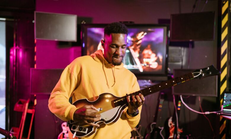 A man in a yellow hoodie plays a guitar simulation game indoors, immersed in gaming fun.