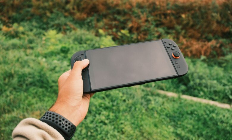 Close-up of a person's hand holding a black handheld gaming console against a green outdoor background.