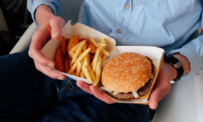 Unrecognizable male with wristwatch in casual clothes sitting on chair and holding box of burger and french fries in hands
