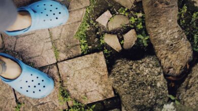 A person wearing blue clogs stands on a stone path surrounded by greenery.