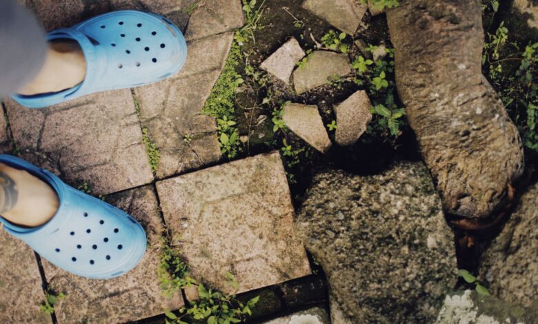 A person wearing blue clogs stands on a stone path surrounded by greenery.