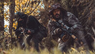Two soldiers camouflaged in a forest, stealthily navigating with rifles in hand.