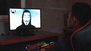 Person using computer in dark room viewing hacker wearing a mask on the screen, symbolizing cyber security threats.