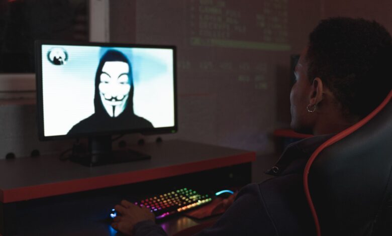 Person using computer in dark room viewing hacker wearing a mask on the screen, symbolizing cyber security threats.