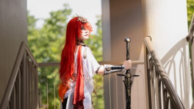 Cosplayer with striking red hair holds a sword on a sunlit stairway, dressed in elaborate costume.
