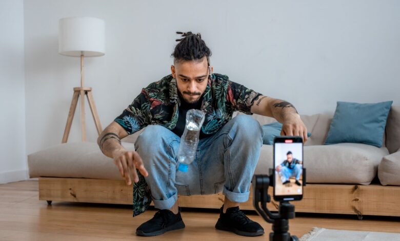 A young man focuses on a bottle flip challenge while filming content at home, capturing it on a smartphone.