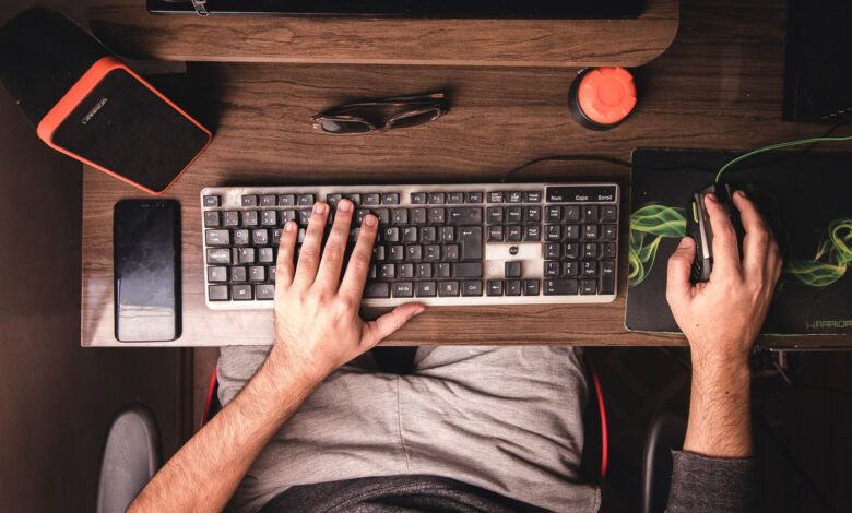 A top-down view of a man using a desktop computer with various electronics on a wooden desk.