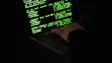 Crop unrecognizable computer geek typing on netbook with codes on screen while hacking system in darkness