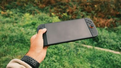 Close-up of a person's hand holding a black handheld gaming console against a green outdoor background.