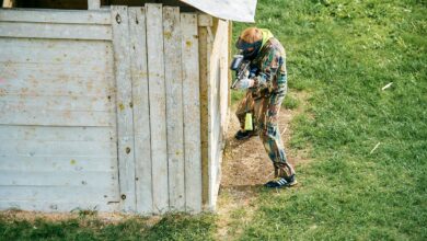 A paintball player taking cover behind a wooden structure on a grassy field during a game.