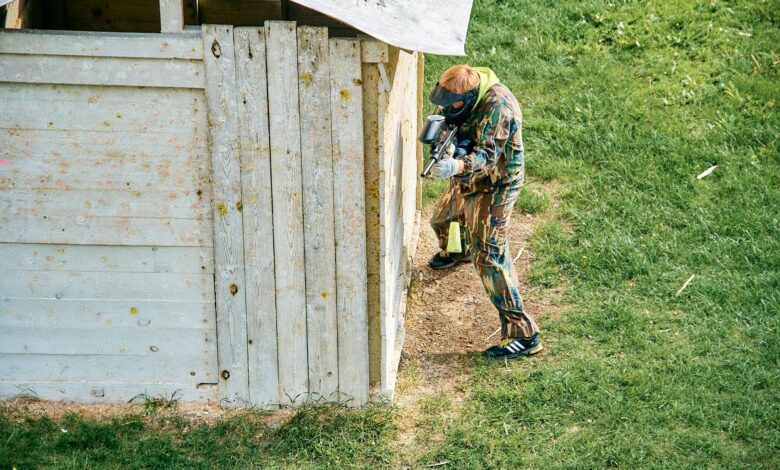 A paintball player taking cover behind a wooden structure on a grassy field during a game.