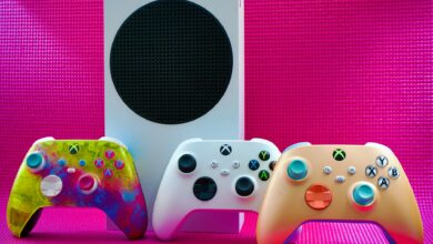 Vibrant gaming controllers displayed with a console on a bold pink backdrop.
