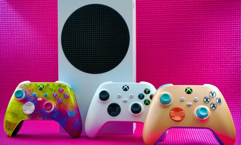 Vibrant gaming controllers displayed with a console on a bold pink backdrop.